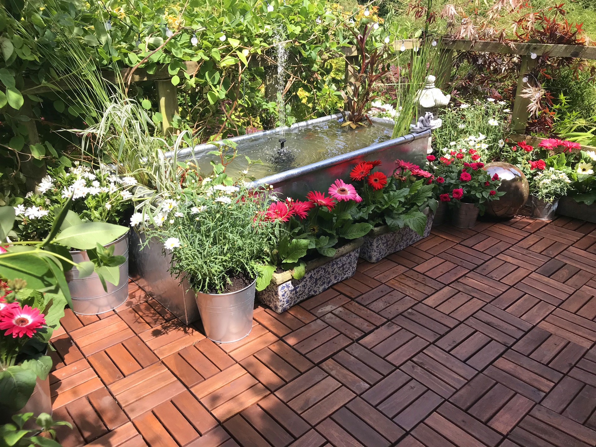 Immagine del balcone della piattaforma della terrazza della casa sull'albero del giardino in estate con giochi d'acqua dello stagno con pompa della fontana solare, pesce rosso, piante marginali, rose in miniatura rosse, fiori di gerbera rosa, piastrelle di